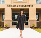 A person wearing a graduation gown and cap stands confidently in front of a building labeled 'Science & Engineering'. The architecture features brickwork and large windows, with a well-maintained pathway and small shrubs leading to the entrance.