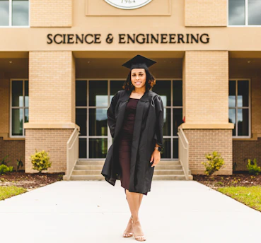 A person wearing a graduation gown and cap stands confidently in front of a building labeled 'Science & Engineering'. The architecture features brickwork and large windows, with a well-maintained pathway and small shrubs leading to the entrance.
