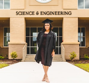 A person wearing a graduation gown and cap stands confidently in front of a building labeled 'Science & Engineering'. The architecture features brickwork and large windows, with a well-maintained pathway and small shrubs leading to the entrance.