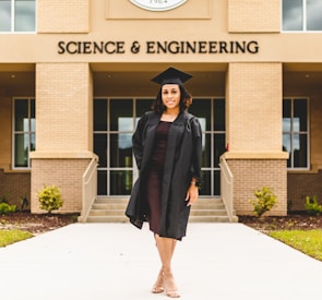 A person wearing a graduation gown and cap stands confidently in front of a building labeled 'Science & Engineering'. The architecture features brickwork and large windows, with a well-maintained pathway and small shrubs leading to the entrance.