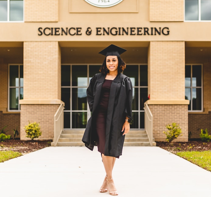 A person wearing a graduation gown and cap stands confidently in front of a building labeled 'Science & Engineering'. The architecture features brickwork and large windows, with a well-maintained pathway and small shrubs leading to the entrance.
