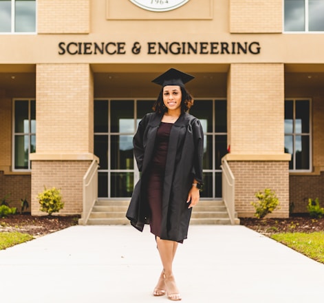 A person wearing a graduation gown and cap stands confidently in front of a building labeled 'Science & Engineering'. The architecture features brickwork and large windows, with a well-maintained pathway and small shrubs leading to the entrance.