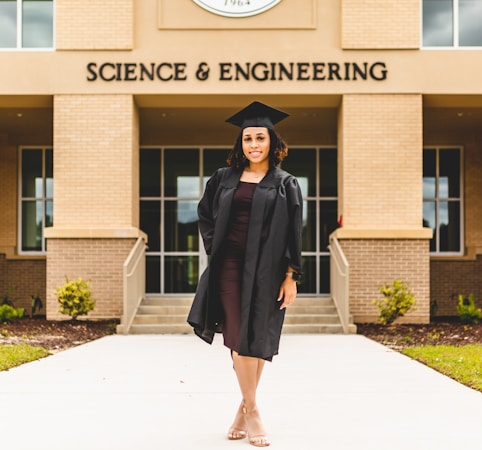 A person wearing a graduation gown and cap stands confidently in front of a building labeled 'Science & Engineering'. The architecture features brickwork and large windows, with a well-maintained pathway and small shrubs leading to the entrance.