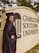 A person in a graduation cap and gown stands beside a sign for Charleston Southern University. The individual is smiling and positioned with one hand on their hip. The surroundings include greenery.