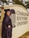 A person in a graduation cap and gown stands beside a sign for Charleston Southern University. The individual is smiling and positioned with one hand on their hip. The surroundings include greenery.