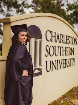 A person in a graduation cap and gown stands beside a sign for Charleston Southern University. The individual is smiling and positioned with one hand on their hip. The surroundings include greenery.