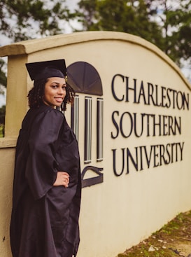 A person in a graduation cap and gown stands beside a sign for Charleston Southern University. The individual is smiling and positioned with one hand on their hip. The surroundings include greenery.