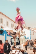 A lively street performance where a performer dressed in a pink costume balances on a hoop above a whimsical, old-fashioned vehicle. Another performer beside the vehicle is in a colorful Victorian-style outfit with face paint and a top hat. The crowd, including both children and adults, watches the performance with interest. The setting is a charming, historical street with white and blue buildings.