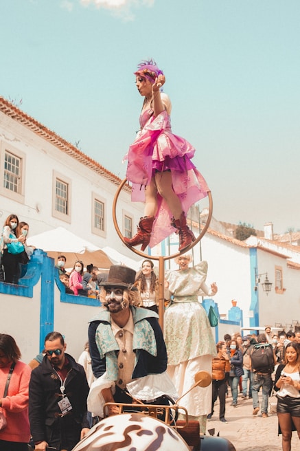 A lively street performance where a performer dressed in a pink costume balances on a hoop above a whimsical, old-fashioned vehicle. Another performer beside the vehicle is in a colorful Victorian-style outfit with face paint and a top hat. The crowd, including both children and adults, watches the performance with interest. The setting is a charming, historical street with white and blue buildings.