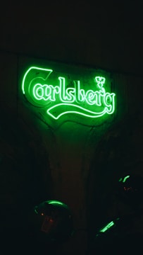 A neon green sign displaying the word 'Carlsberg' is illuminated against a dark background. The glow of the sign casts a bright light, highlighting its unique typography. Below the sign, partially visible objects in the dim light might include parts of a helmet or a motorcycle.