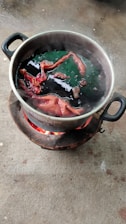 A metal pot placed on an outdoor stove with flaming charcoal. Inside the pot, pieces of fabric are immersed in a dark liquid, possibly undergoing a dyeing process. Steam rises from the pot, suggesting the liquid is boiling.