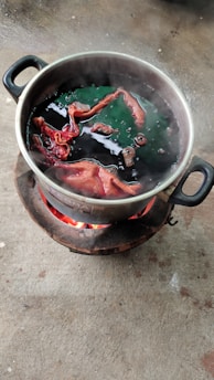 A metal pot placed on an outdoor stove with flaming charcoal. Inside the pot, pieces of fabric are immersed in a dark liquid, possibly undergoing a dyeing process. Steam rises from the pot, suggesting the liquid is boiling.