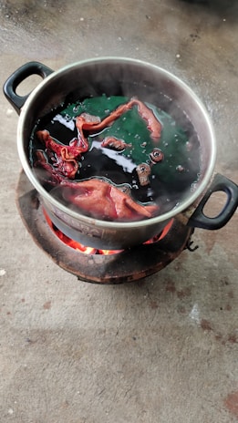 A metal pot placed on an outdoor stove with flaming charcoal. Inside the pot, pieces of fabric are immersed in a dark liquid, possibly undergoing a dyeing process. Steam rises from the pot, suggesting the liquid is boiling.