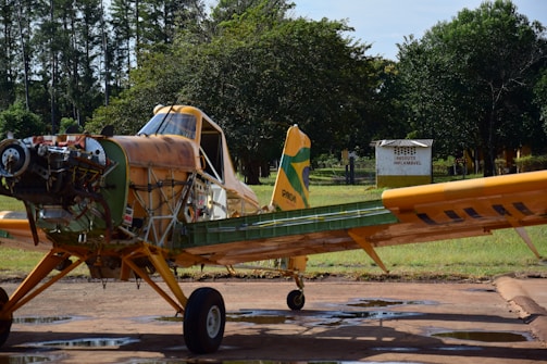 An agricultural aircraft is parked on a tarmac with a disassembled front section, exposing its engine and various components. The aircraft is painted in shades of yellow and green, and it sits against a backdrop of trees and grass. A sign in the background indicates the presence of a flammable product.