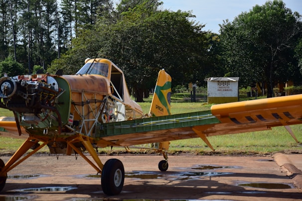 An agricultural aircraft is parked on a tarmac with a disassembled front section, exposing its engine and various components. The aircraft is painted in shades of yellow and green, and it sits against a backdrop of trees and grass. A sign in the background indicates the presence of a flammable product.