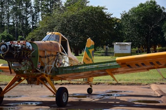 An agricultural aircraft is parked on a tarmac with a disassembled front section, exposing its engine and various components. The aircraft is painted in shades of yellow and green, and it sits against a backdrop of trees and grass. A sign in the background indicates the presence of a flammable product.