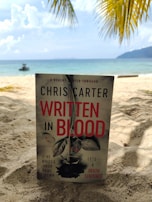 A book titled 'Written in Blood' by Chris Carter stands upright on a sandy beach. The background features the sea with gentle waves and a distant boat on the water. Overhead, palm fronds frame the scene, suggesting a tropical setting. The sky is partly cloudy.