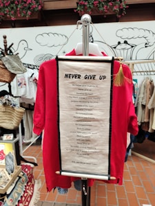 A red shirt displayed on a hanger with an inspirational scroll attached featuring a motivational quote by the Dalai Lama. The background includes decorative elements, various items, and hanging baskets in a shop setting.