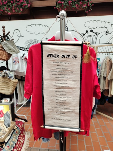 A red shirt displayed on a hanger with an inspirational scroll attached featuring a motivational quote by the Dalai Lama. The background includes decorative elements, various items, and hanging baskets in a shop setting.