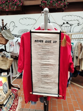 A red shirt displayed on a hanger with an inspirational scroll attached featuring a motivational quote by the Dalai Lama. The background includes decorative elements, various items, and hanging baskets in a shop setting.