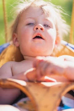 A young child with light hair appears in a close-up shot, capturing a moment of curiosity or wonder. The background is blurred with hints of green, suggesting an outdoor setting, perhaps at a park.