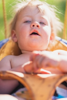 A young child with light hair appears in a close-up shot, capturing a moment of curiosity or wonder. The background is blurred with hints of green, suggesting an outdoor setting, perhaps at a park.