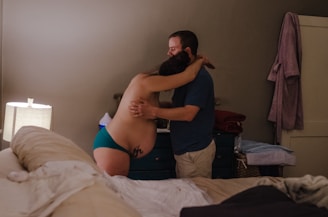 A pregnant woman wearing teal underwear is embracing a man in a blue shirt in a bedroom setting. The room is softly lit by a lamp on a bedside table, and there is a neatly made bed in the foreground. Towels are stacked on a dresser, and a bathrobe hangs on a door.