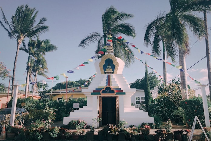 A white stupa-like structure with a golden top, adorned with colorful prayer flags, is surrounded by lush tropical plants and tall palm trees. The sky is clear and blue, creating a serene and peaceful atmosphere. A small garden with various potted plants and flowering bushes enhances the tranquil setting.