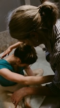 Two people appear to be engaged in a soothing, intimate moment, possibly involving support or care. One person is in a tub of water, with their head resting on their arms, while the other person leans over from outside the tub, gently touching and comforting them.