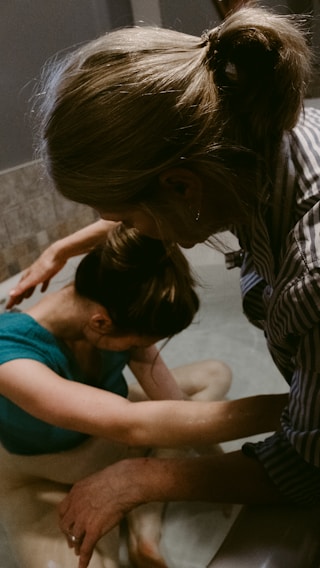 Two people appear to be engaged in a soothing, intimate moment, possibly involving support or care. One person is in a tub of water, with their head resting on their arms, while the other person leans over from outside the tub, gently touching and comforting them.