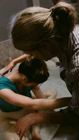 Two people appear to be engaged in a soothing, intimate moment, possibly involving support or care. One person is in a tub of water, with their head resting on their arms, while the other person leans over from outside the tub, gently touching and comforting them.
