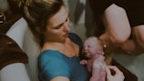 A woman holds a newborn baby close to her chest while sitting in a bathtub. The baby is freshly born, with a visible amount of vernix on the skin. The scene suggests an intimate and tender moment between the mother and child during or right after a water birth.