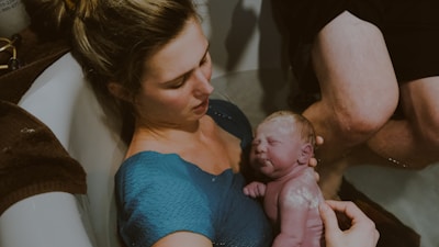 A woman holds a newborn baby close to her chest while sitting in a bathtub. The baby is freshly born, with a visible amount of vernix on the skin. The scene suggests an intimate and tender moment between the mother and child during or right after a water birth.
