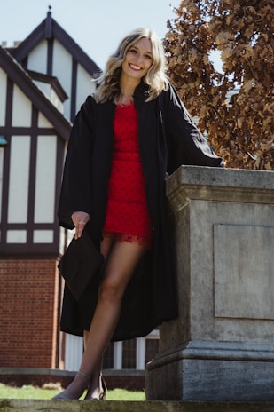 A person wearing a red dress under a black graduation gown stands confidently on a stone platform outdoors, with a building featuring Tudor-style architecture in the background. The person is holding a graduation cap and smiling, with blonde hair cascading over their shoulders. There is a tree with browned leaves beside them.