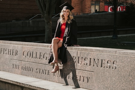 A person in a graduation cap and gown is sitting on a stone ledge with engraved text reading 'Fisher College of Business, The Ohio State University'. The setting appears to be outdoors with a brick building and trees in the background.
