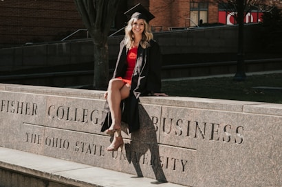 A person in a graduation cap and gown is sitting on a stone ledge with engraved text reading 'Fisher College of Business, The Ohio State University'. The setting appears to be outdoors with a brick building and trees in the background.