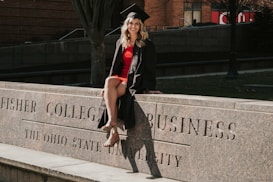 A person in a graduation cap and gown is sitting on a stone ledge with engraved text reading 'Fisher College of Business, The Ohio State University'. The setting appears to be outdoors with a brick building and trees in the background.
