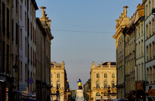 A symmetrical view of a historic European square with ornate, classical architecture lining both sides. In the center stands a statue draped with a yellow and blue cloth, surrounded by sunlight casting a warm tone on the buildings. The sky is clear.