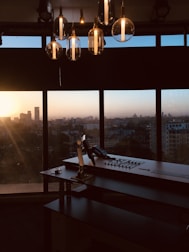 An office setup with a large desk and a control panel in front of a panoramic window. The city skyline is visible in the background, blanketed by the warm hues of a setting sun. Hanging overhead are several modern, glass pendant lights reflecting the ambient light.