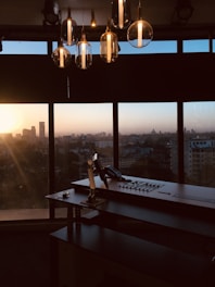 An office setup with a large desk and a control panel in front of a panoramic window. The city skyline is visible in the background, blanketed by the warm hues of a setting sun. Hanging overhead are several modern, glass pendant lights reflecting the ambient light.