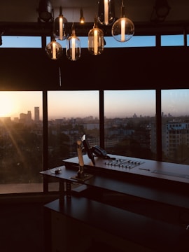 An office setup with a large desk and a control panel in front of a panoramic window. The city skyline is visible in the background, blanketed by the warm hues of a setting sun. Hanging overhead are several modern, glass pendant lights reflecting the ambient light.