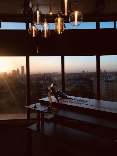 An office setup with a large desk and a control panel in front of a panoramic window. The city skyline is visible in the background, blanketed by the warm hues of a setting sun. Hanging overhead are several modern, glass pendant lights reflecting the ambient light.