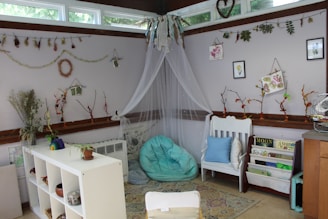 A cozy and artistic corner is adorned with natural elements and craft items. There is a white chair with cushions and a bean bag in soft blue tones. The walls are decorated with various hanging crafts made from twigs, leaves, and flowers. A white shelf holds plants and small items, while books and art are displayed nearby. The overall setting suggests a nature-inspired, creative space with soft lighting streaming through large windows.