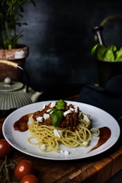 A plate of spaghetti topped with a rich meat sauce, garnished with basil leaves and crumbled cheese, is set on a wooden table. The background features a blurred dark setting with hints of a plant and other kitchen elements.