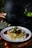 A plate of spaghetti topped with a rich meat sauce, garnished with basil leaves and crumbled cheese, is set on a wooden table. The background features a blurred dark setting with hints of a plant and other kitchen elements.