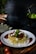 A plate of spaghetti topped with a rich meat sauce, garnished with basil leaves and crumbled cheese, is set on a wooden table. The background features a blurred dark setting with hints of a plant and other kitchen elements.