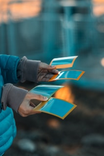 Hands holding several photographs with a blurred background. The photos have a blue tint and are framed with an orange border.