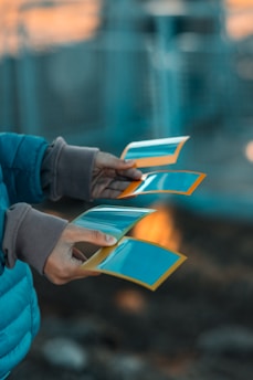 Hands holding several photographs with a blurred background. The photos have a blue tint and are framed with an orange border.