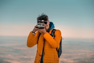 A person wearing an orange jacket and backpack is holding a vintage instant camera up to their eye, taking a photograph against a backdrop of a serene sky.