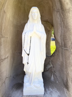 A white statue of a figure with hands in prayer holding a black rosary is situated within a stone alcove. The statue features a serene expression and wears a draped gown. The light creates a soft shadow effect around the figure.